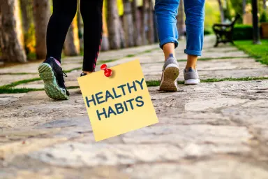 Afbeelding van twee vrouwen die samen wandelen op een parkpad, met een notitie met de tekst 'Healthy Habits' vastgeprikt op de voorgrond, symboliserend een frisse start en gezonde gewoonten voor 2025.