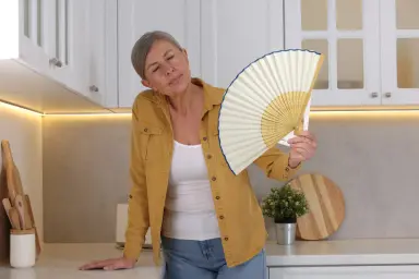 Vrouw in de overgang met grijs haar, die een waaier vasthoudt terwijl ze zich zichtbaar ongemakkelijk voelt, waarschijnlijk door opvliegers, in een moderne keuken.